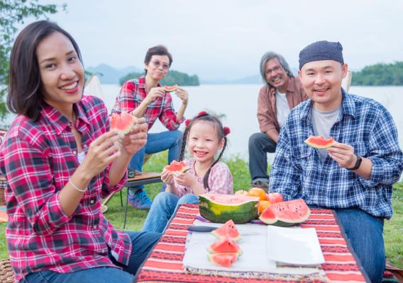 A family eating watermelon outside at a camping site near a lake. A mother, father, and their young daughter are sitting at a table, with two grandparents sitting closer to the lake in the background