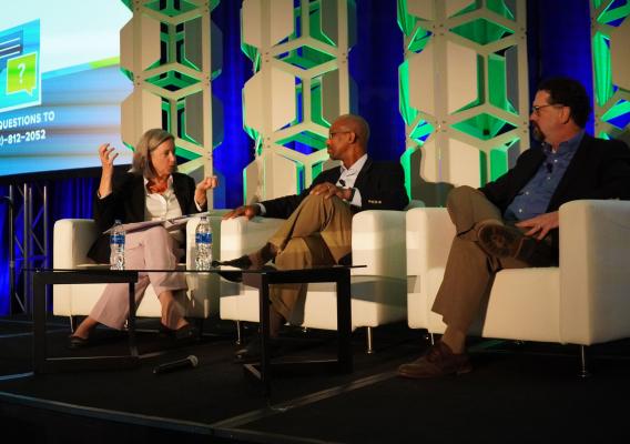 A panel of speakers in professional attire sit in three white chairs lined up on a stage with a coffee table and water bottles in front of them