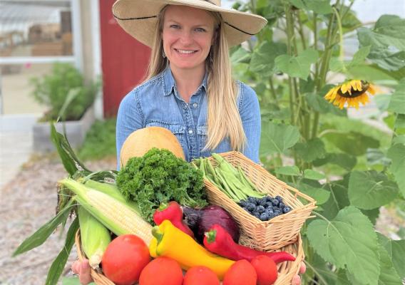 Amanda Sweetman wearing a wide-brimmed straw hat holding a basket of vegetables with sunflowers in the background